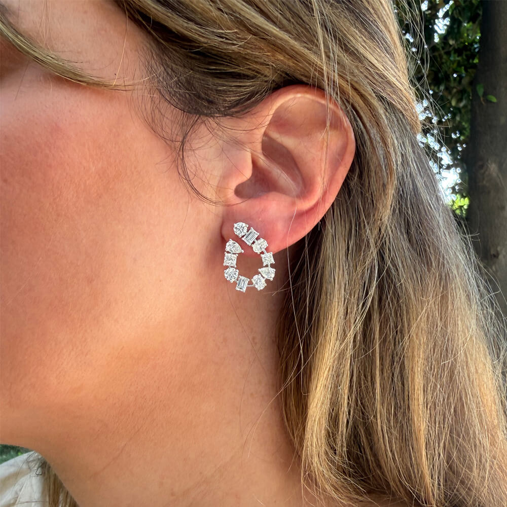A close-up of a woman's ear wearing an 18K white gold Estate circular dangle earring adorned with multiple clear mixed shape diamonds, set against her hair and a blurred outdoor background.