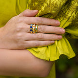 Close-up of a woman wearing three rings with green, blue, and diamond gemstones on a yellow fabric background.