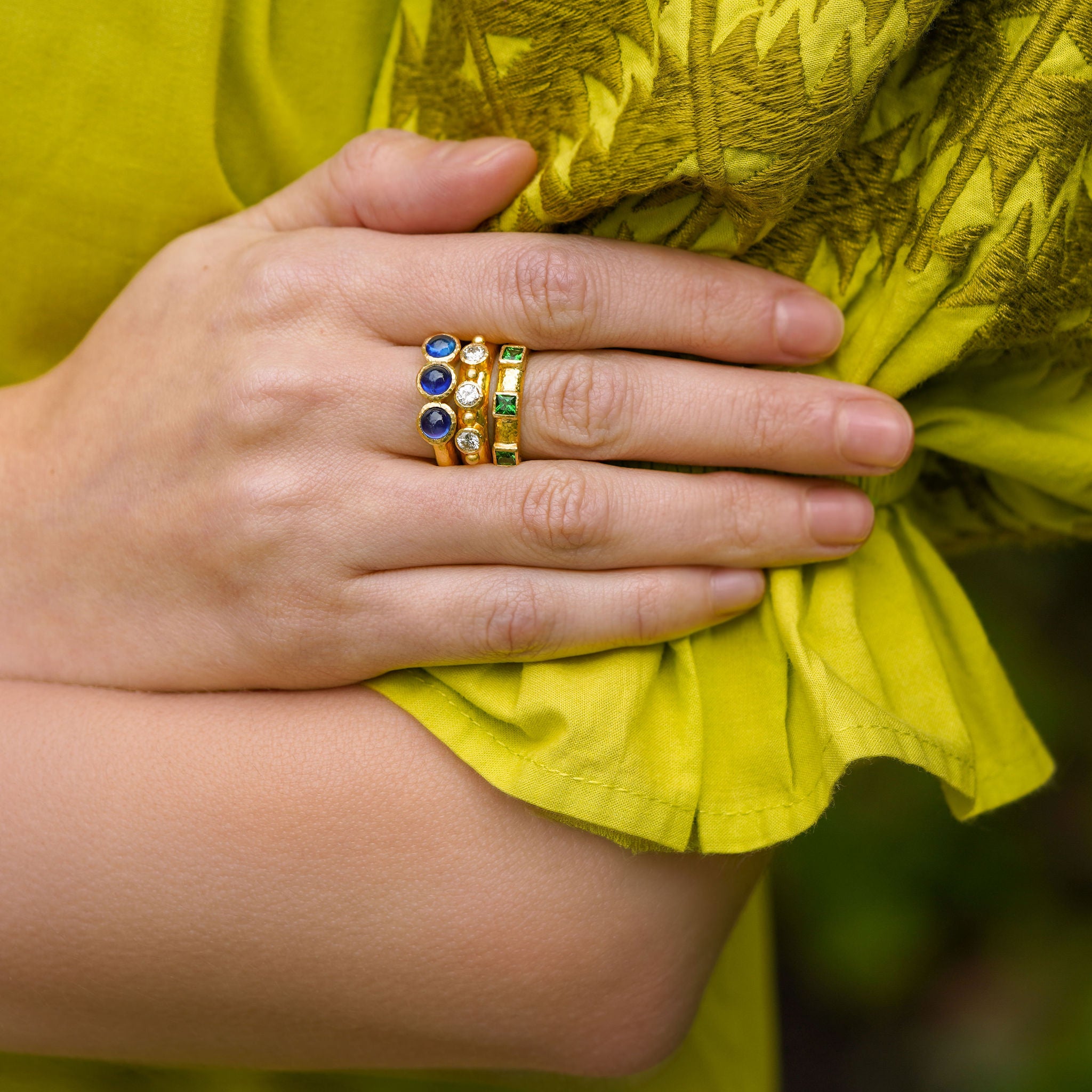 Close-up of a woman wearing three rings with green, blue, and diamond gemstones on a yellow fabric background.