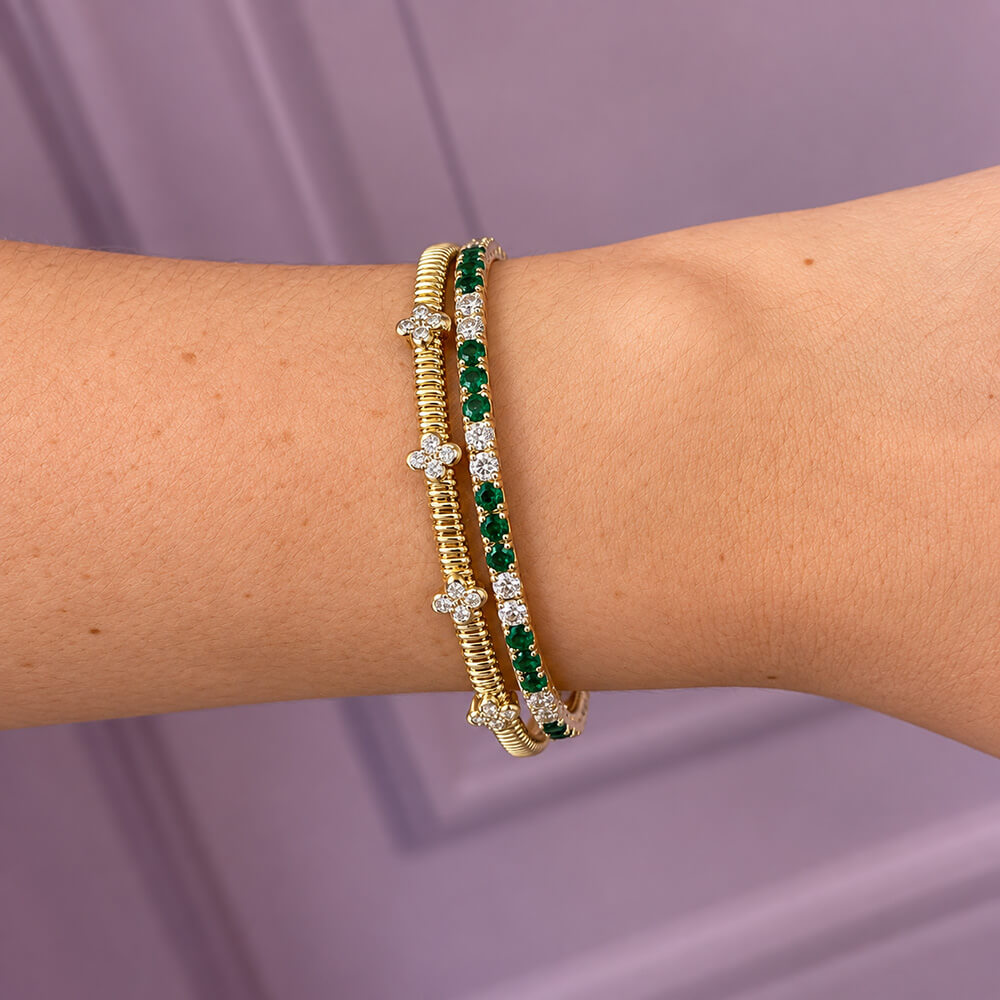 A close-up of a woman's wrist wearing two elegant bracelets: one yellow gold bracelet with diamond clover stations, and one yellow gold bracelet with green emeralds and clear diamonds, against a soft lavender background.