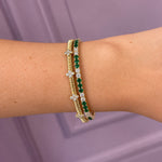 A close-up of a woman's wrist wearing two elegant bracelets: one yellow gold bracelet with diamond clover stations, and one yellow gold bracelet with green emeralds and clear diamonds, against a soft lavender background.
