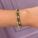 A close-up of a woman's wrist wearing two elegant bracelets: one yellow gold bracelet with diamond clover stations, and one yellow gold bracelet with green emeralds and clear diamonds, against a soft lavender background.