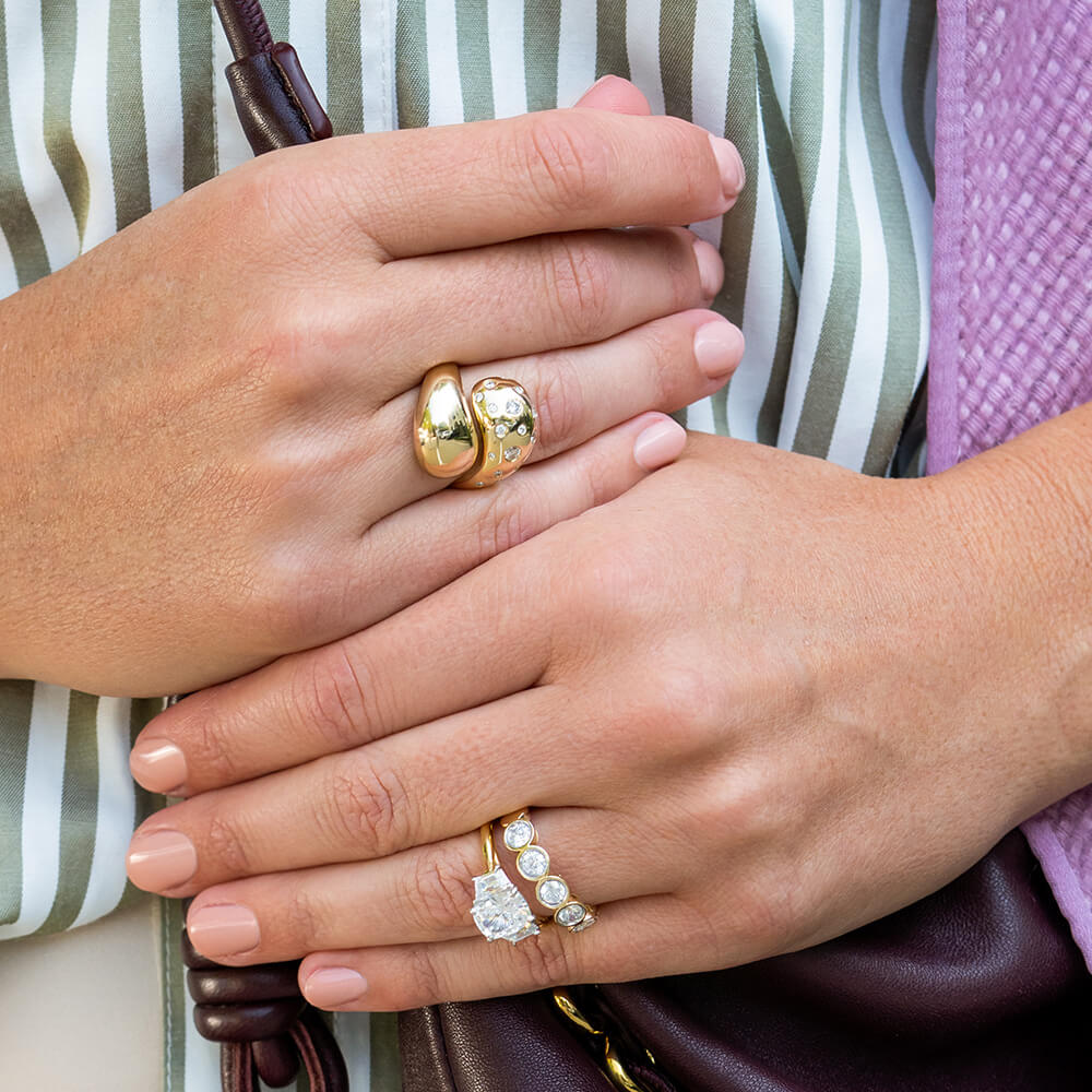 Close-up of two hands wearing 18K yellow gold rings, including a chunky gold ring with scattered diamonds, and another 18k gold ring with clear diamonds, worn over a green and white striped top.