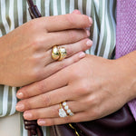Close-up of two hands wearing 18K yellow gold rings, including a chunky gold ring with scattered diamonds, and another 18k gold ring with clear diamonds, worn over a green and white striped top.