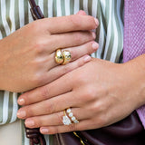 Close-up of two hands wearing 18K yellow gold rings, including a chunky gold ring with scattered diamonds, and another 18k gold ring with clear diamonds, worn over a green and white striped top.