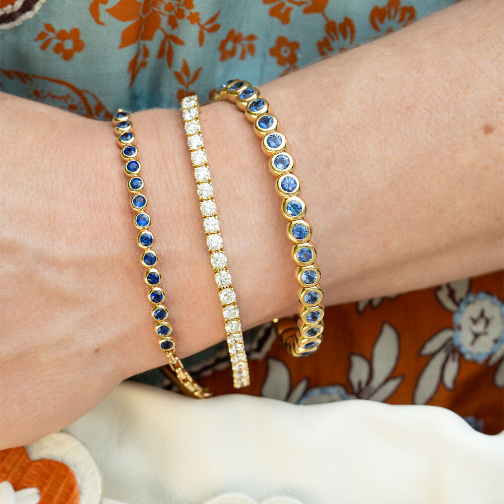 Three tennis-style bracelets on a woman's wrist: two 18K yellow gold bracelets with blue sapphire stones and one 18K yellow gold bracelet with clear diamonds, against a floral garment background.