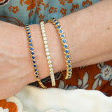 Three tennis-style bracelets on a woman's wrist: two 18K yellow gold bracelets with blue sapphire stones and one 18K yellow gold bracelet with clear diamonds, against a floral garment background.