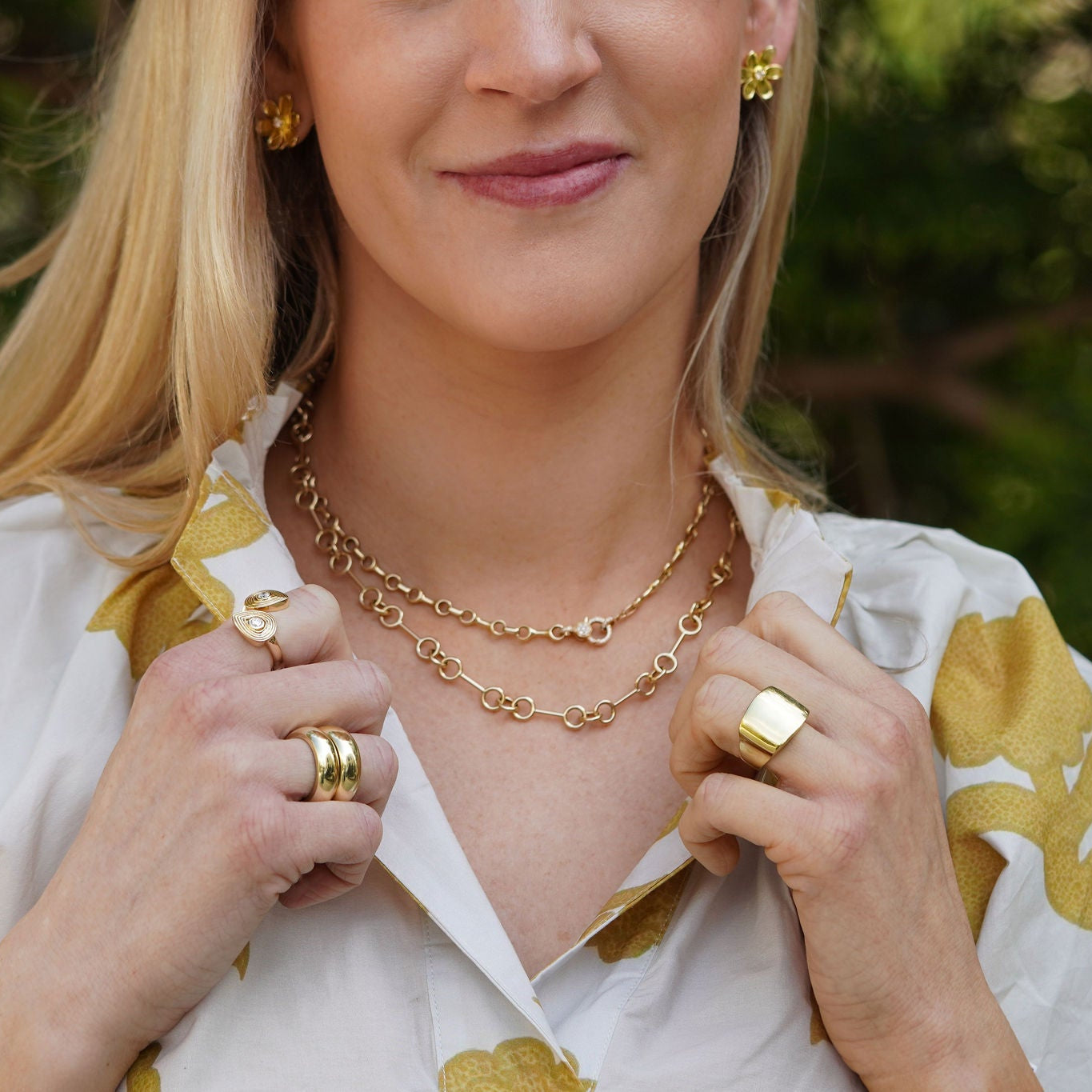 Woman wearing gold jewelry outdoors with greenery in the background