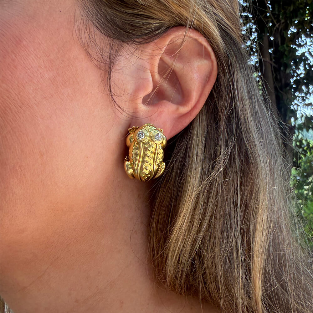 A close-up of a woman's ear wearing an 18K yellow gold frog-shaped earring with small clear diamonds.