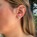 A close-up of a woman's ear wearing a small 18K yellow gold butterfly-shaped stud earring with clear diamonds, set against an outdoor background with blurred greenery.