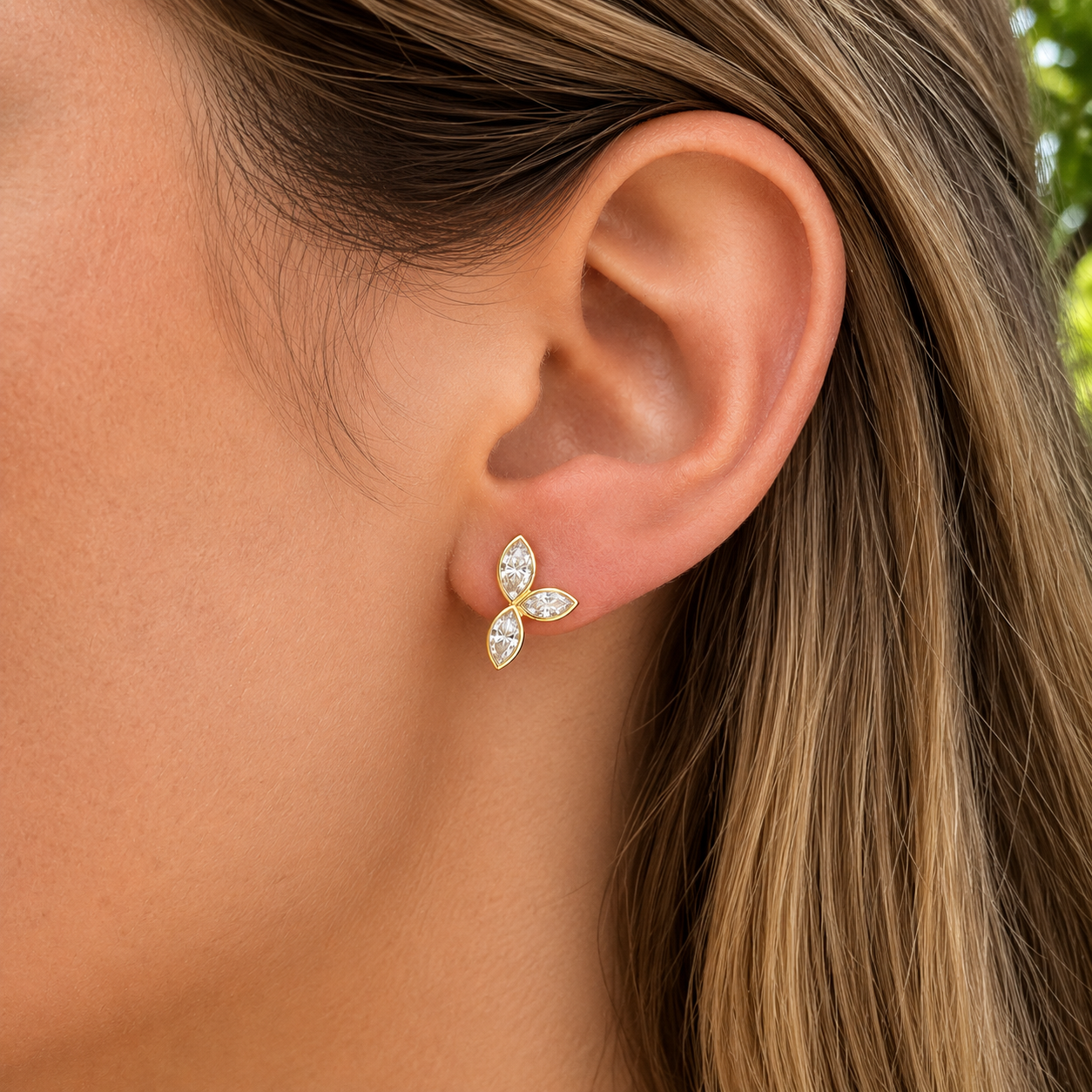A close-up of a woman's ear wearing a small gold stud earring with three marquise-cut clear stones, set against her hair and skin.