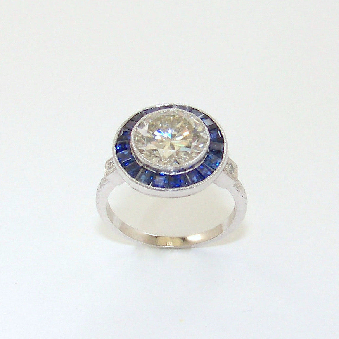 A platinum ring with a central large round diamond stone surrounded by smaller sapphire insets, set on a plain white background.