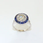 A platinum ring with a central large round diamond stone surrounded by smaller sapphire insets, set on a plain white background.