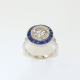 A platinum ring with a central large round diamond stone surrounded by smaller sapphire insets, set on a plain white background.